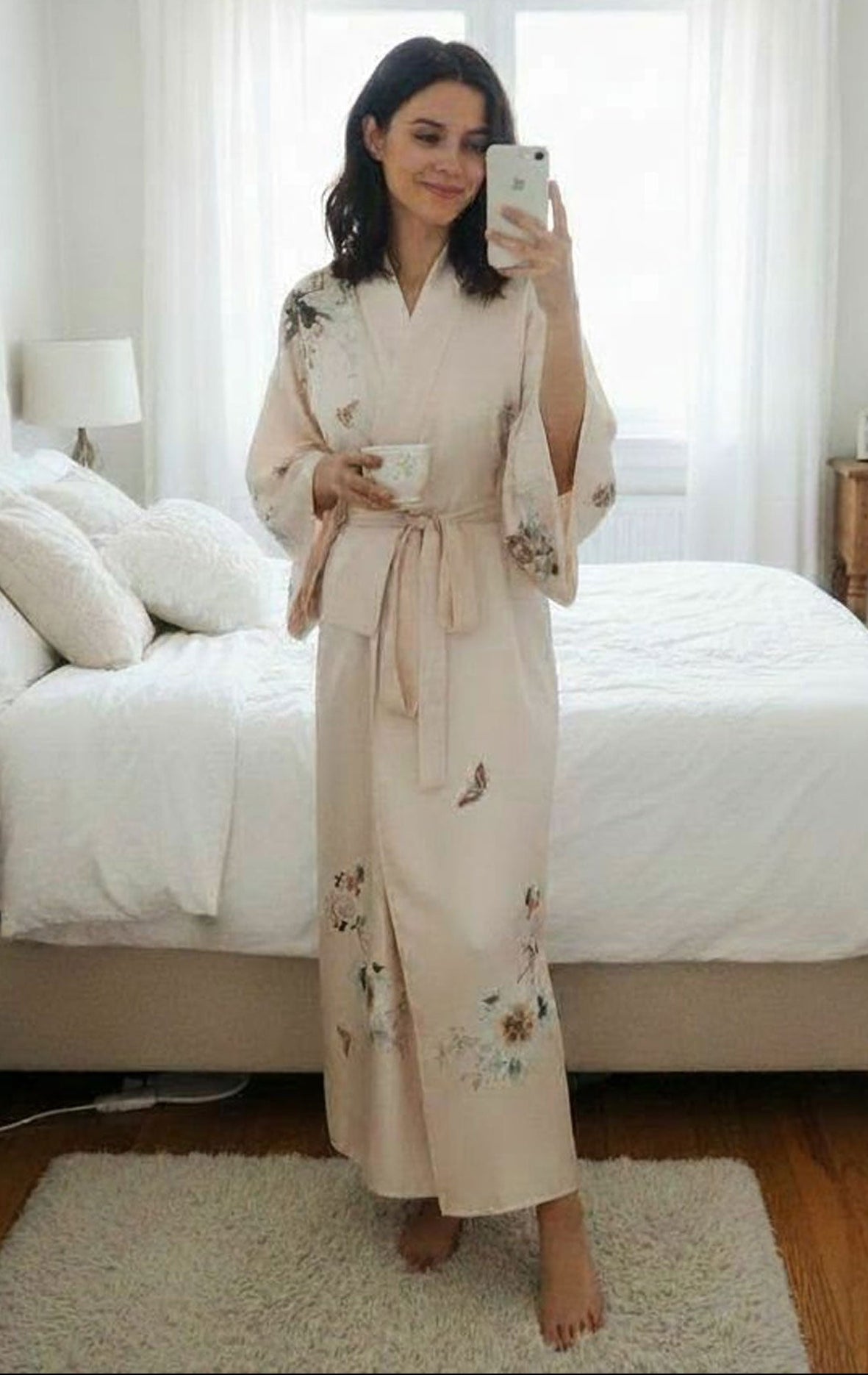 Woman in a floral robe taking a mirror selfie in a bedroom.