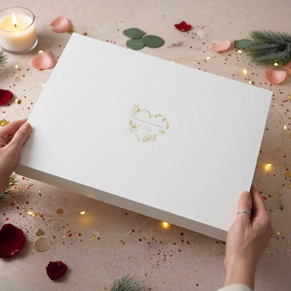 White box with a logo held by hands on a decorative surface with candles and flowers.