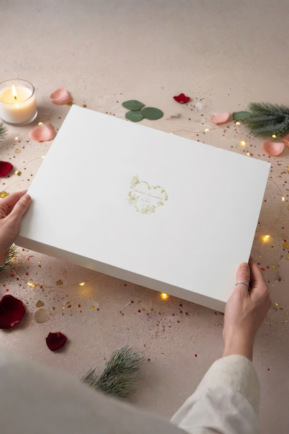 White gift box with a gold logo held by hands on a decorative surface with candles and rose petals.
