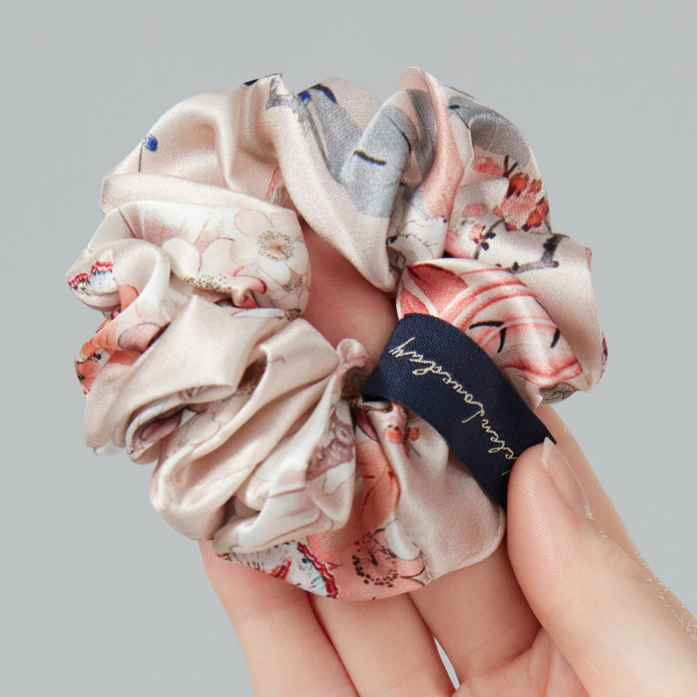 Hand holding a floral scrunchie with a brand label against a gray background