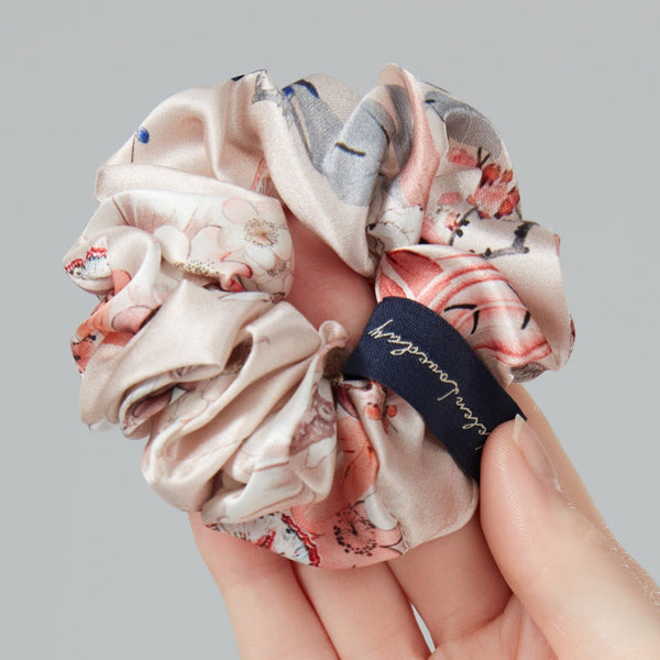 Hand holding a floral scrunchie with a brand label against a gray background